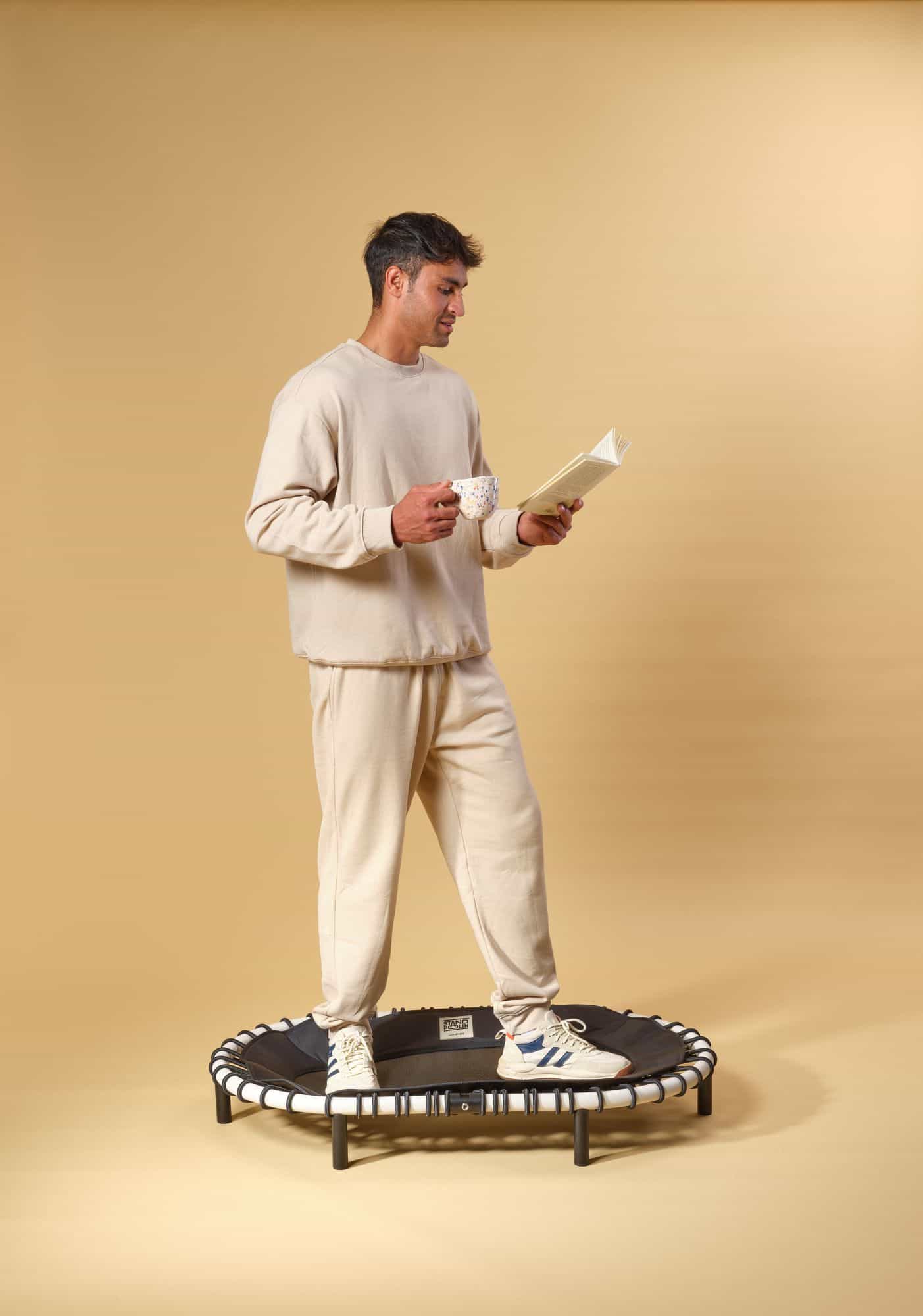 Mann sitzt entspannt auf einem Standpolin mit cremefarbenem Rahmen, schwarzen Gummiseilen und sichtbarem Standpolin Logo und liest ein Buch mit einer Tasse Kaffee in der Hand vor gelbem Hintergrund
