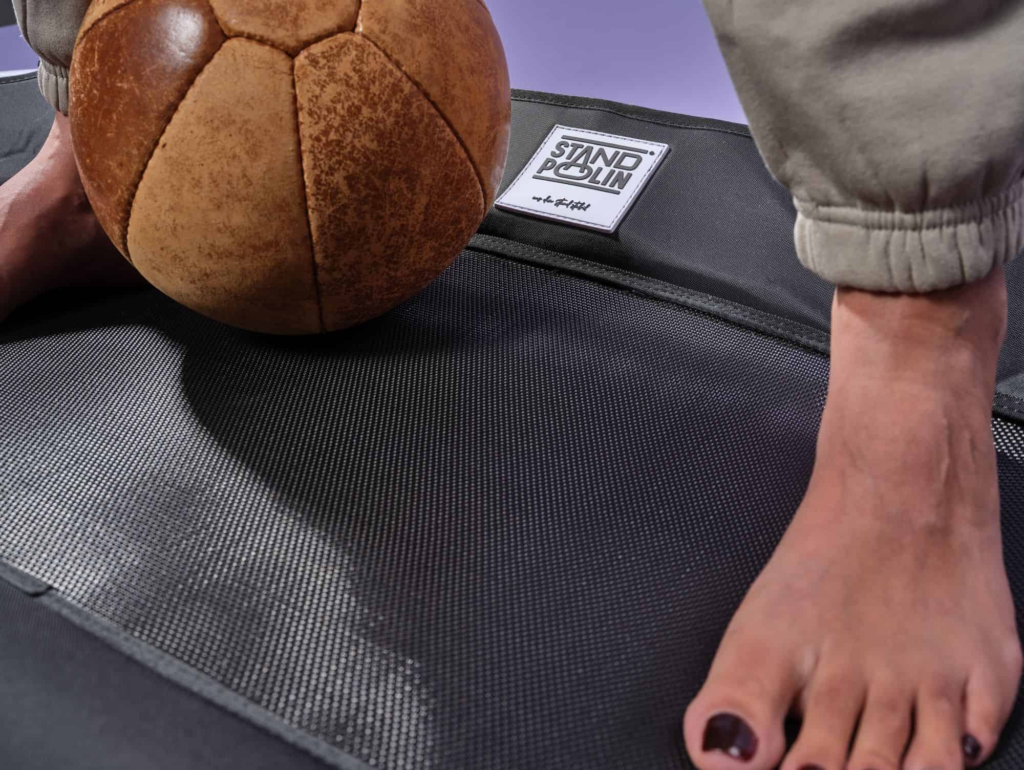 Nahaufnahme eines Standpolin mit schwarzer Sprungfläche, sichtbarem Standpolin Logo, einem braunen Lederball und einem Fuß auf dem Trampolin