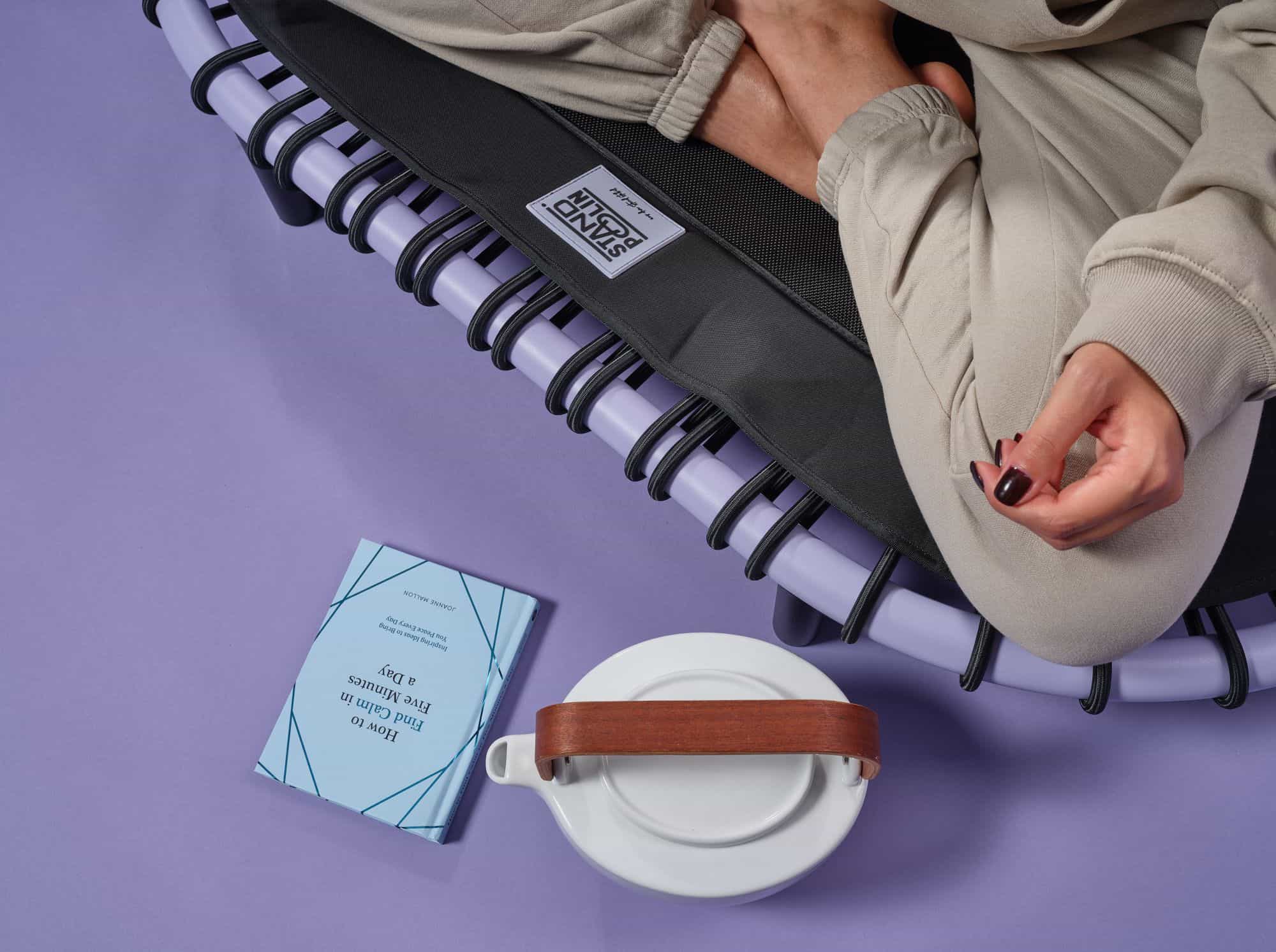 Person sitzt entspannt auf einem Standpolin mit lilafarbenem Rahmen, schwarzen Gummiseilen und sichtbarem Standpolin Logo, daneben liegen ein Buch und eine Tasse auf violettem Hintergrund