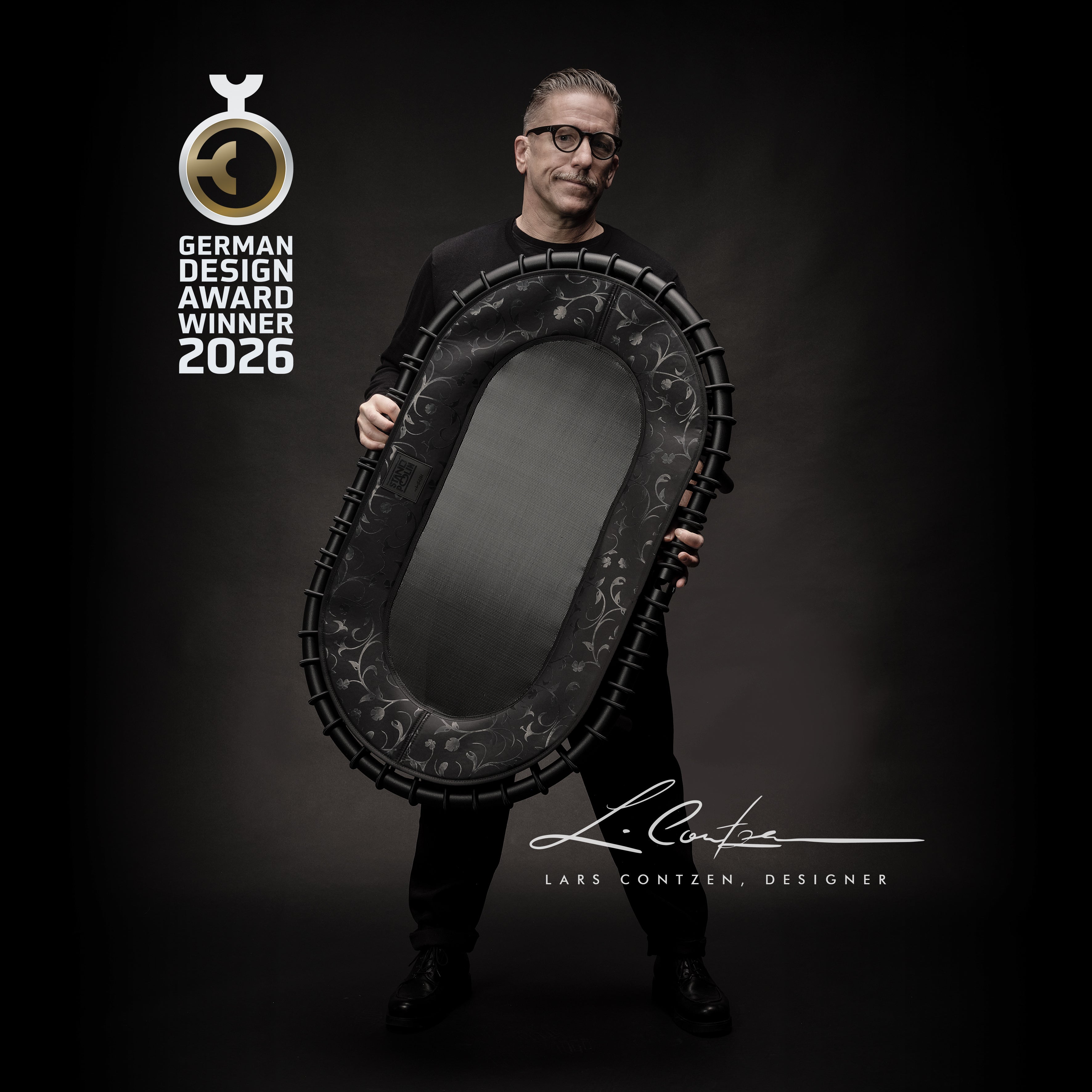 Der Designer Lars Contzen hält die Standpolin Contzen Edition in Anthrazit mit German Design Award Winner 2026 Auszeichnung, floraler Randgestaltung und dunkler Sprungfläche.