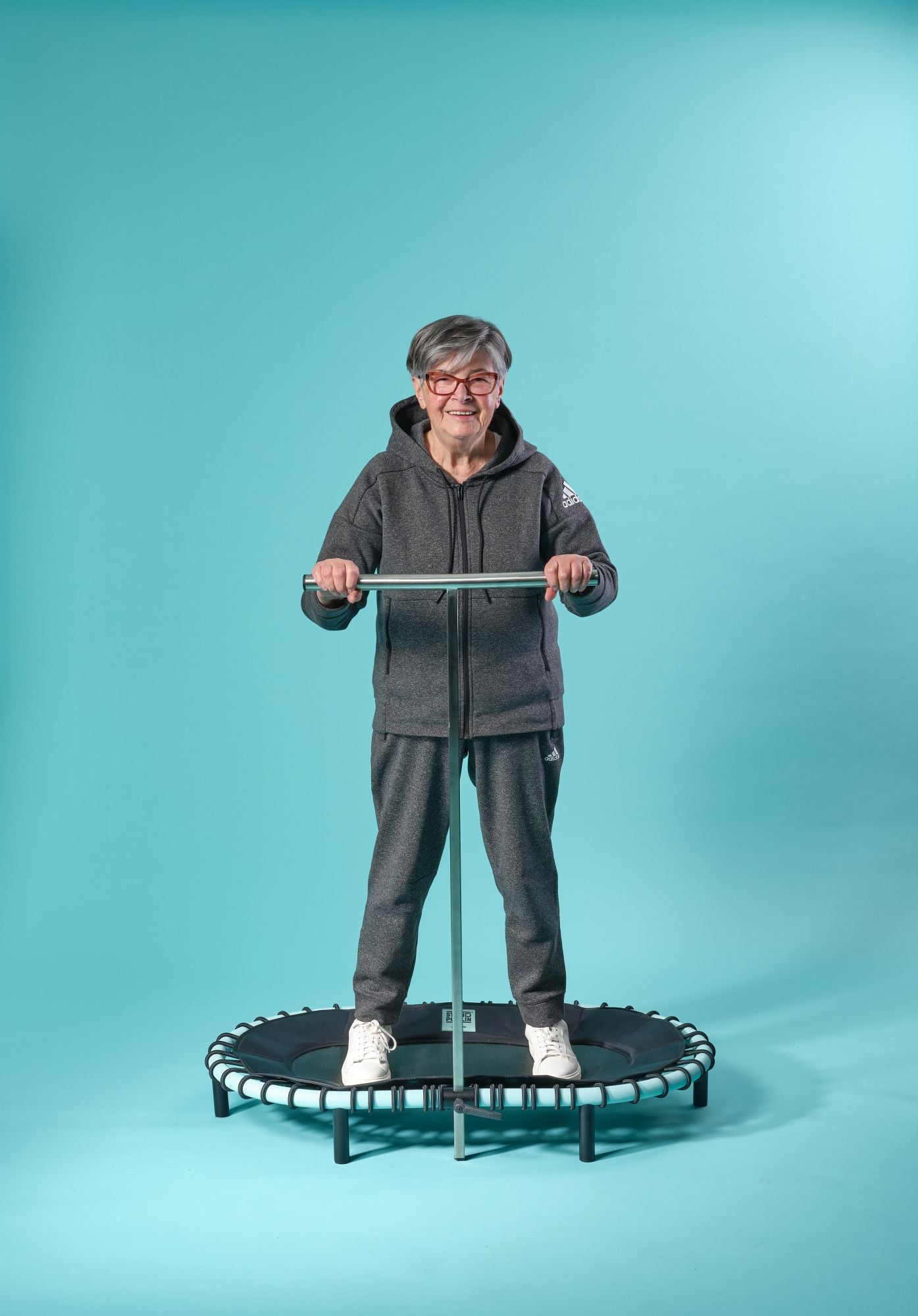 Seniorin steht auf einem Standpolin mit Aqua Rahmen, hält sich an der Haltestange fest und trainiert sicher vor türkisfarbenem Studiohintergrund.