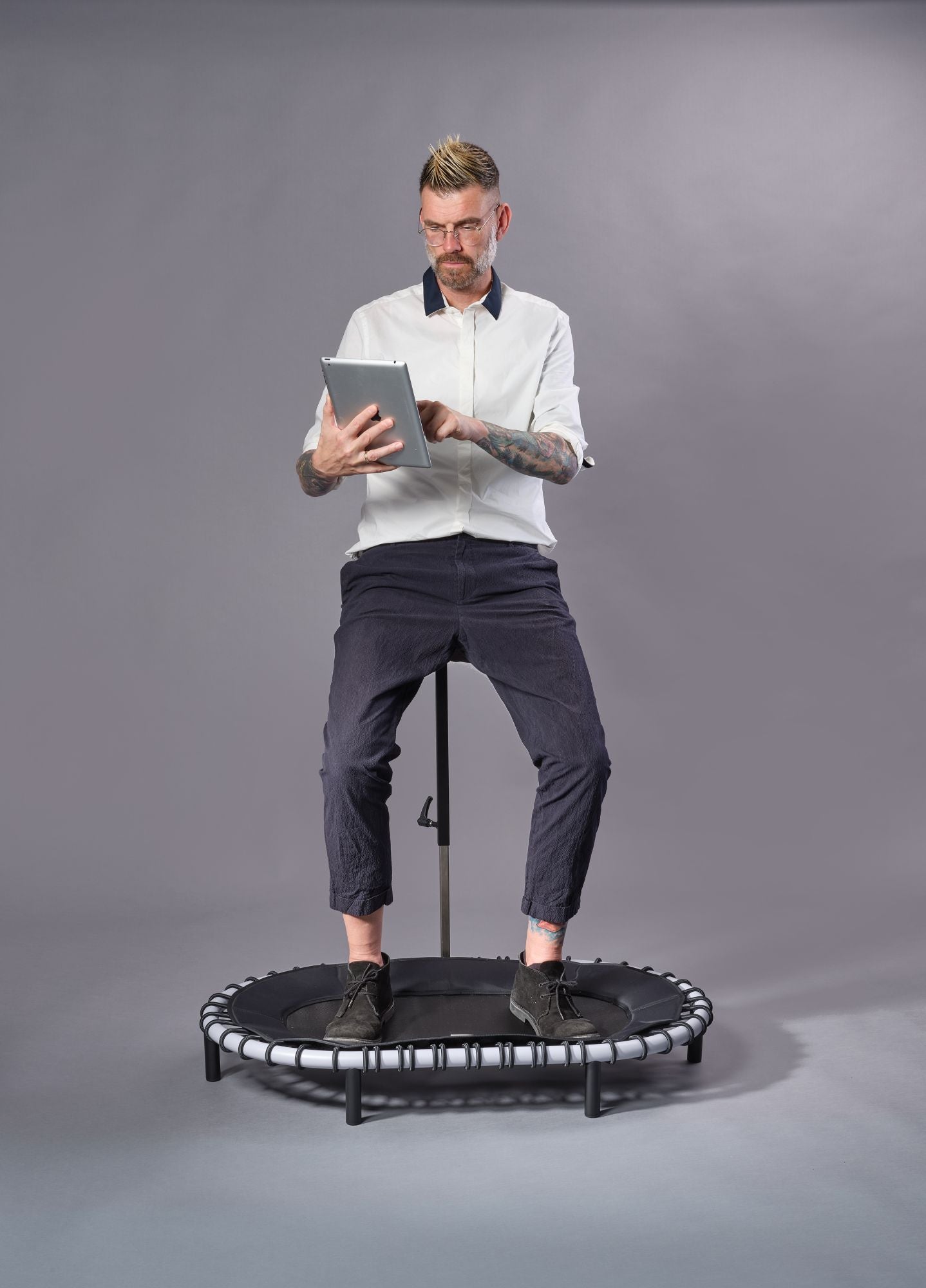 Mann sitzt auf einem Standpolin mit Hellgrau Rahmen, nutzt ein Tablet und kombiniert ergonomisches Arbeiten mit Bewegung vor grauem Studiohintergrund.