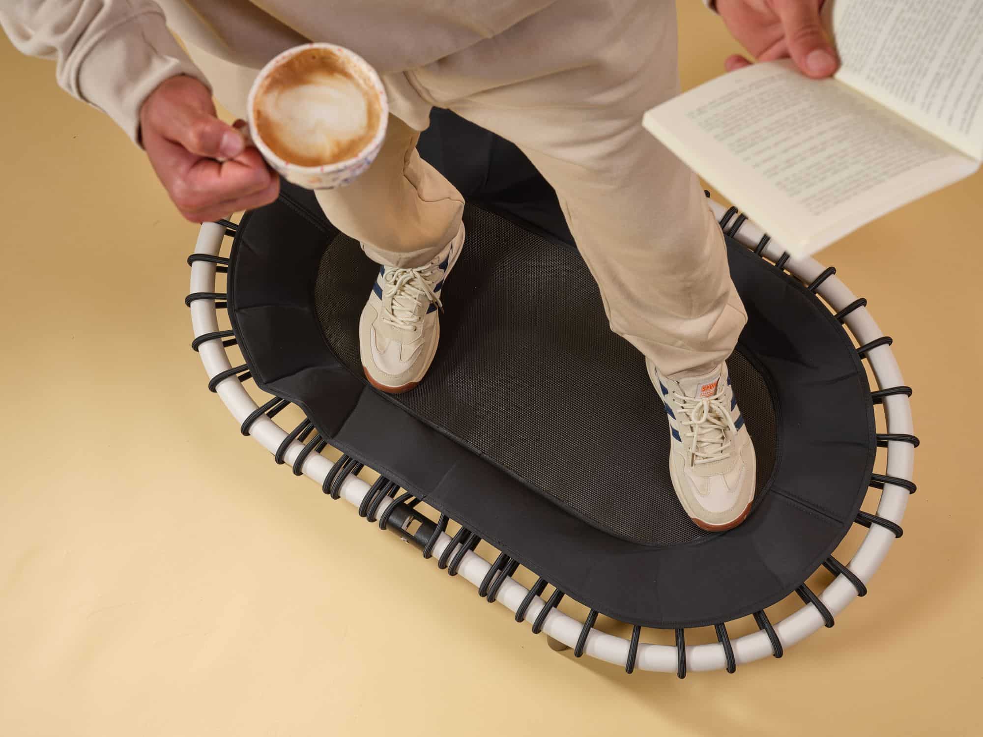 Draufsicht auf eine Person, die auf einem Standpolin mit cremefarbenem Rahmen und schwarzen Gummiseilen steht, dabei eine Tasse Kaffee hält und ein Buch liest vor gelbem HintergrundDraufsicht auf eine Person, die auf einem Standpolin mit cremefarbenem Rahmen und schwarzen Gummiseilen steht, dabei eine Tasse Kaffee hält und ein Buch liest vor gelbem Hintergrund