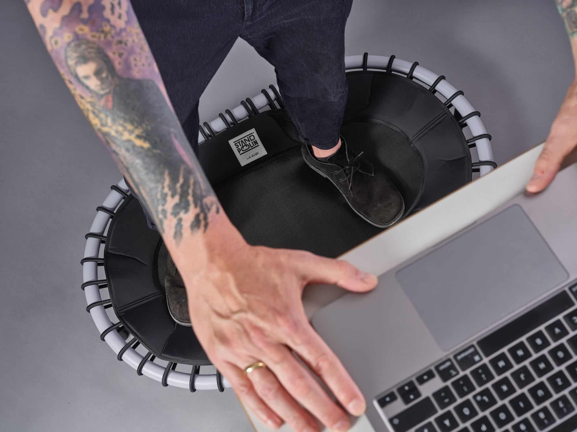Mann arbeitet an einem Laptop und steht dabei auf einem Standpolin mit silbernem Rahmen, schwarzen Gummiseilen und sichtbarem Standpolin Logo vor grauem Hintergrund