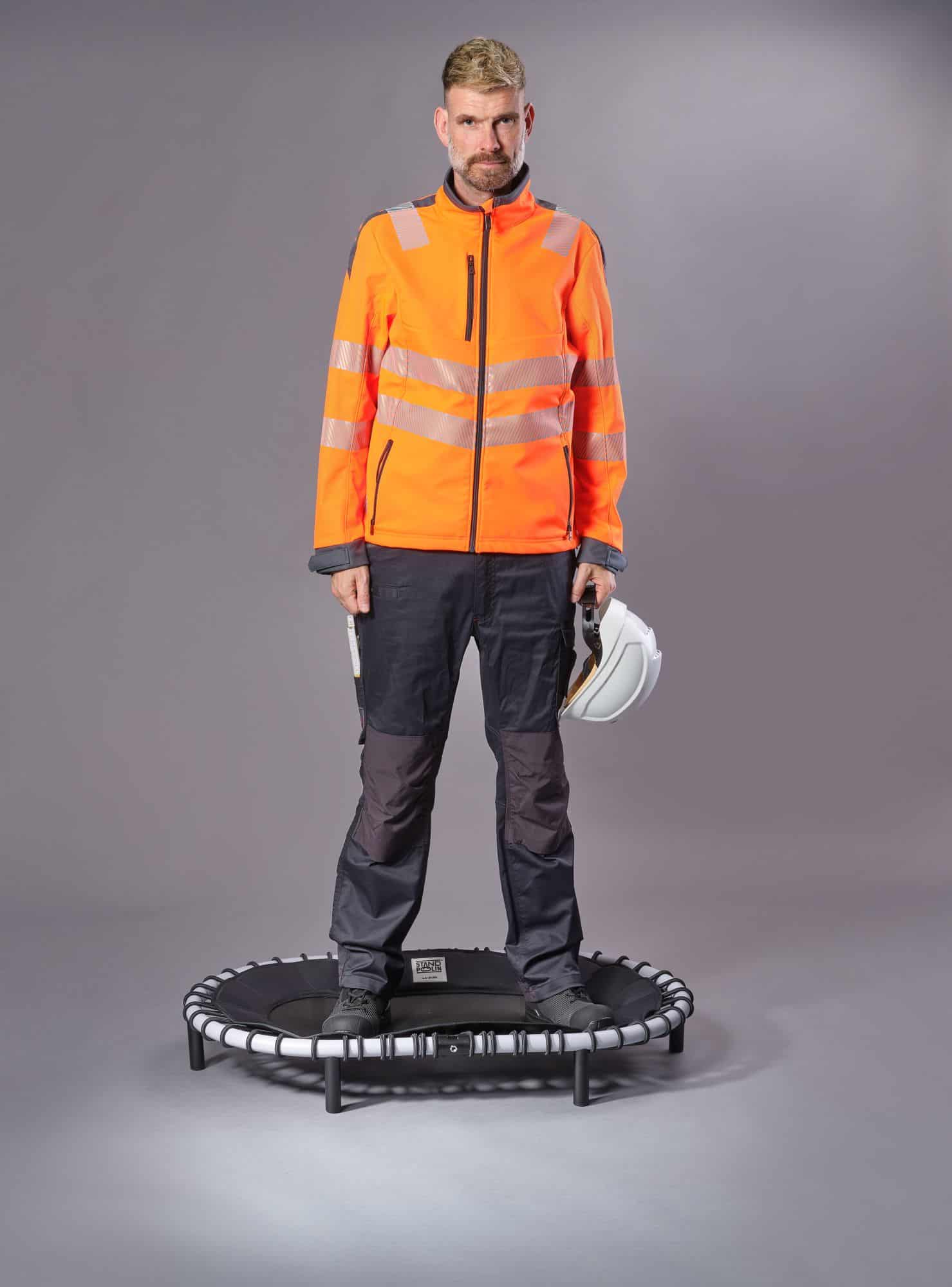 Mann in orangefarbener Warnschutzjacke steht mit weißem Schutzhelm in der Hand auf einem Standpolin mit schwarzem Bezug und hellgrauem Rahmen vor grauem Studiohintergrund